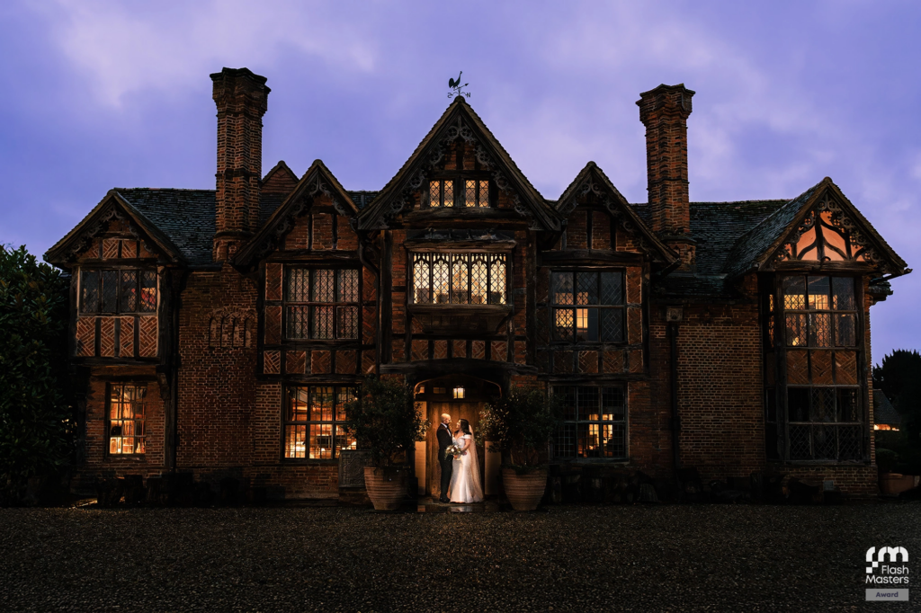 A bride and groom stand in front of a large, illuminated, Tudor-style manor at dusk, holding hands under the main entrance, with warm light glowing from the windows.