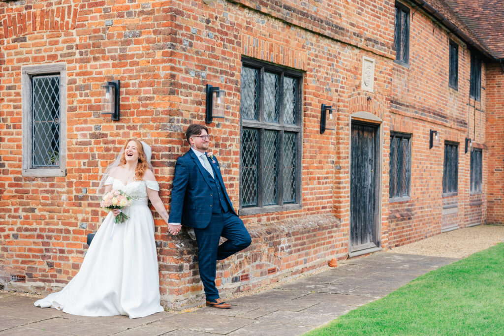 A bride and groom stand back-to-back on either side of a brick building, holding hands round the corner. The bride smiles and holds a bouquet, while the groom leans against the wall, both dressed in wedding attire.