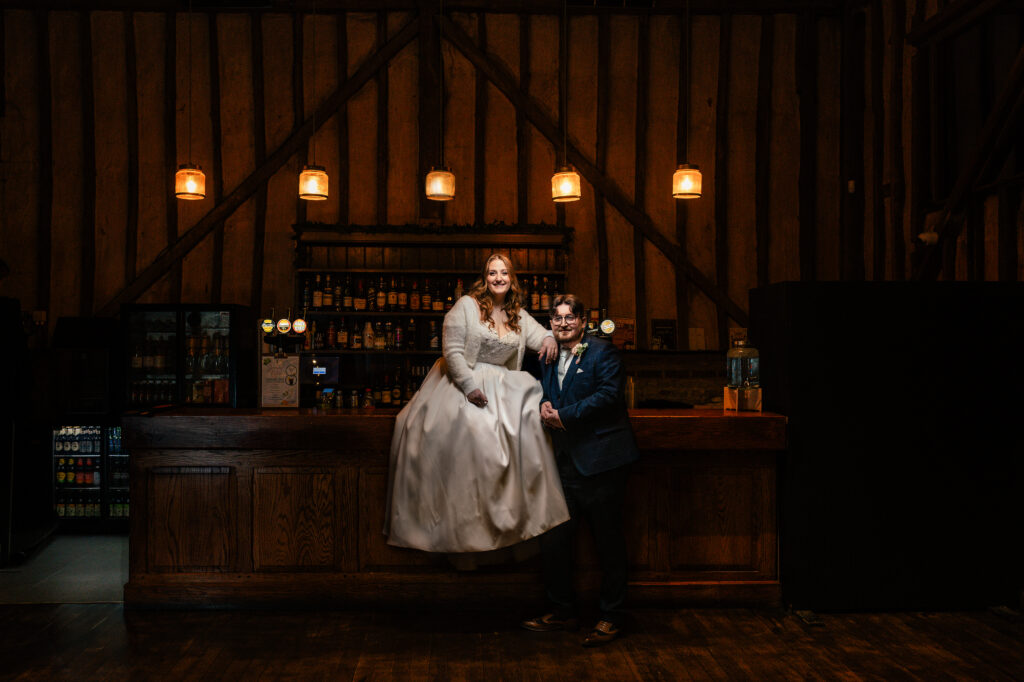 A bride in a white dress sits on a wooden bar, smiling, while a groom in a blue suit stands beside her, holding her hand. The bar is warmly lit with hanging lights and has shelves of bottles behind it.