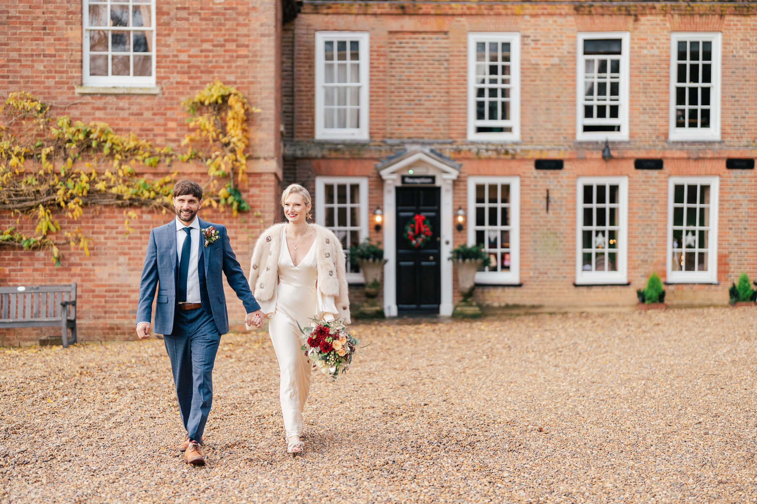 A bride in a white dress and fur coat holds a bouquet and walks hand-in-hand with a groom in a blue suit outside a large brick building with many windows and a black door.