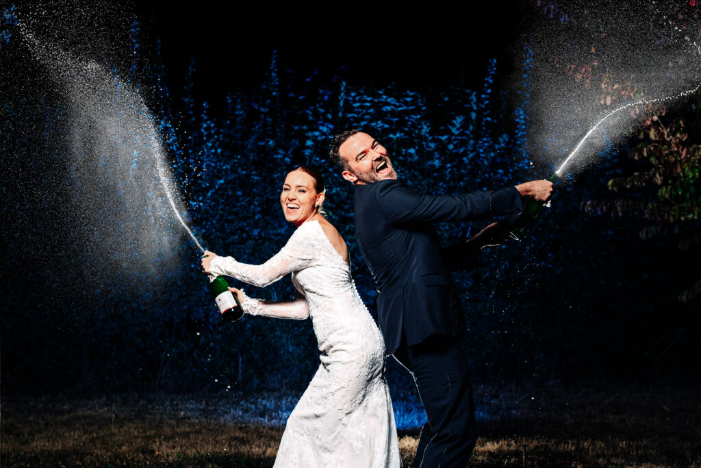 A bride in a white dress and a groom in a dark suit stand back-to-back at night, both laughing and spraying champagne into the air, creating a festive and joyful scene.