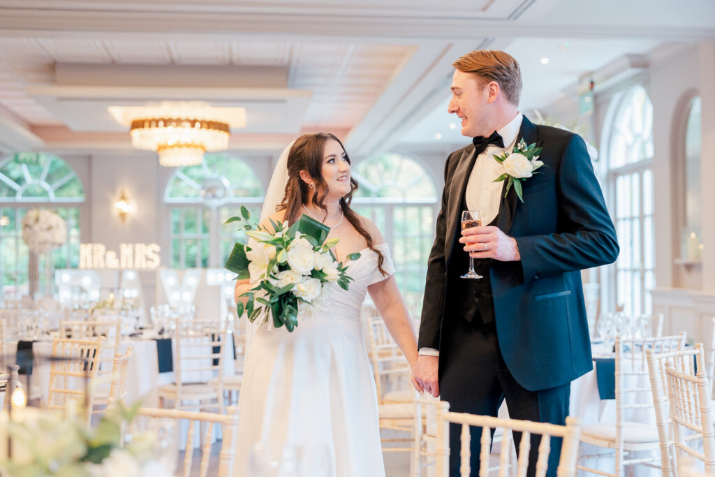 A bride and groom smile at each other whilst holding hands in an elegant, bright wedding venue. The bride holds a white bouquet and the groom holds a drink. Décor includes white flowers and chairs, with a “Mr & Mrs” sign in the background.