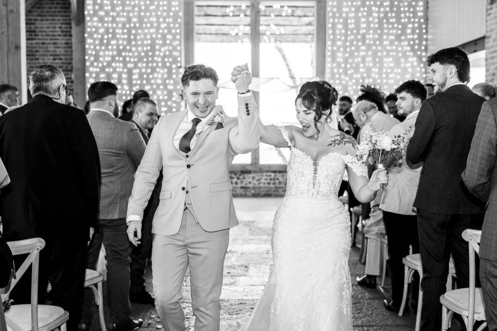 A joyful bride and groom walk down the aisle, holding hands and celebrating, surrounded by smiling guests in a decorated venue with fairy lights in the background.