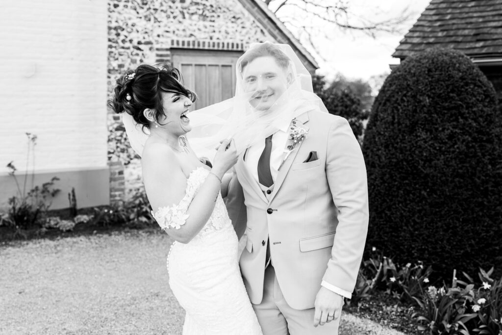 A bride and groom laugh together outside; the bride playfully covers the groom’s face with her veil. The groom wears a suit, and they stand in front of a stone building and garden.