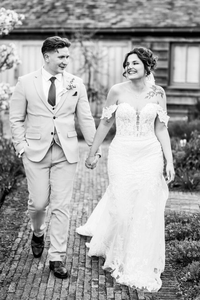 A smiling bride and groom walk hand-in-hand outdoors, both dressed in wedding attire. The bride wears a lace gown; the groom wears a light-coloured suit and tie. They look at each other, happy and joyful, with a rustic building behind them.