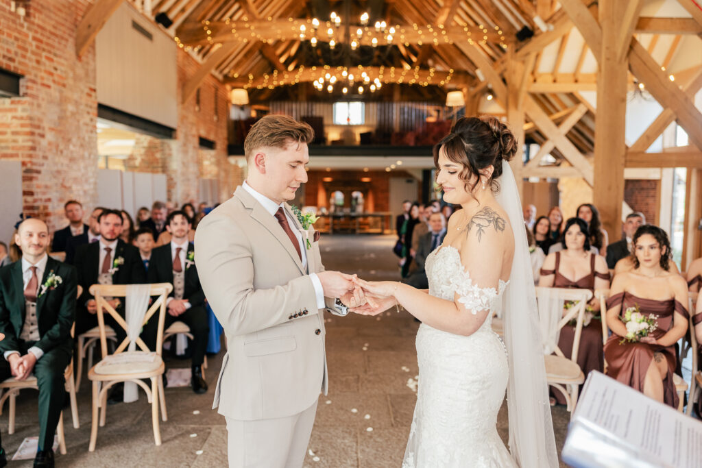 A groom in a light suit puts a ring on the bride’s finger during a wedding ceremony in a rustic, timber-beamed venue, with guests seated and watching attentively in the background.