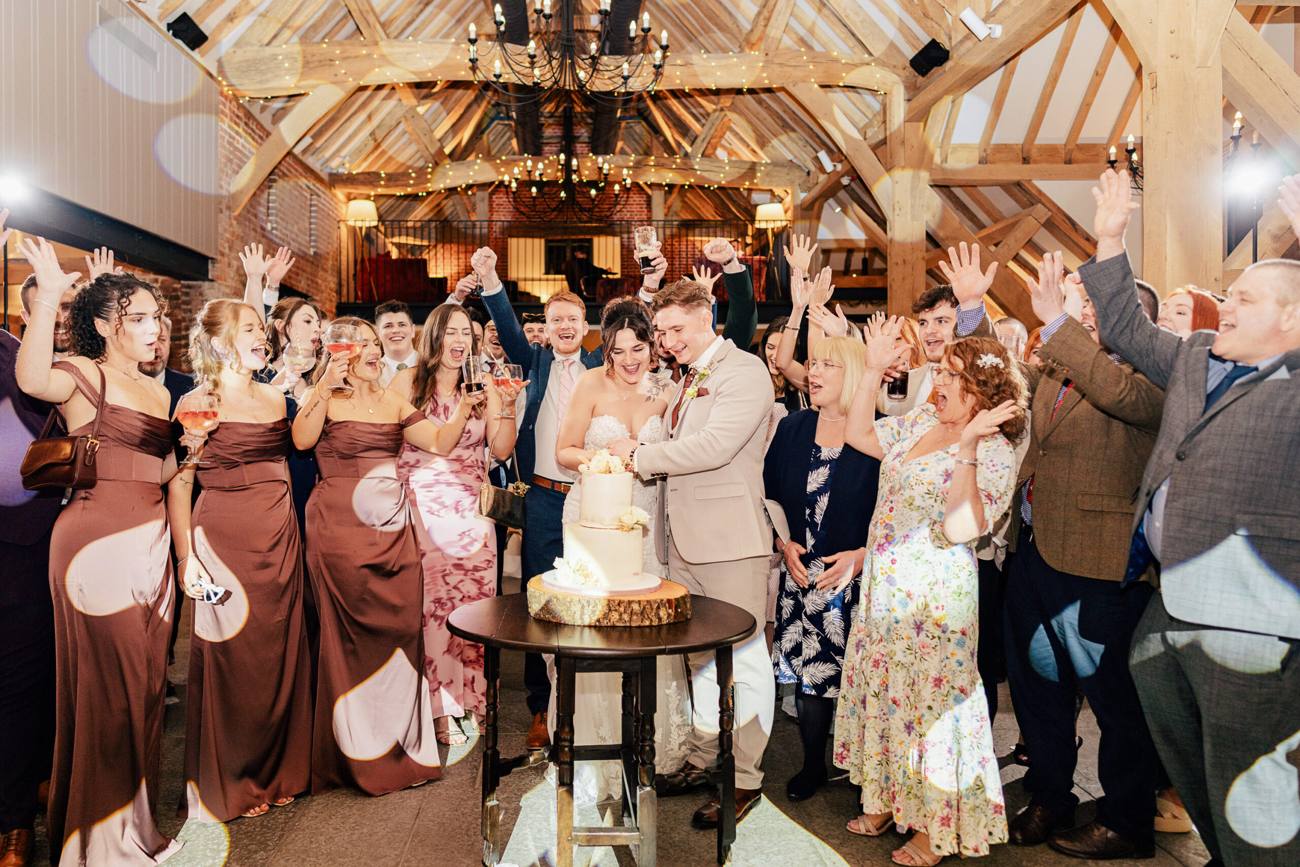A newlywed couple cuts their wedding cake, surrounded by cheering guests in formal attire inside a rustic barn venue with wooden beams and warm lighting.
