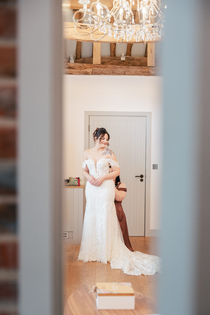 A bride in a white off-shoulder gown smiles as another woman helps adjust her dress, seen through a doorway. A chandelier hangs above, and a box sits on the wooden floor in the foreground.