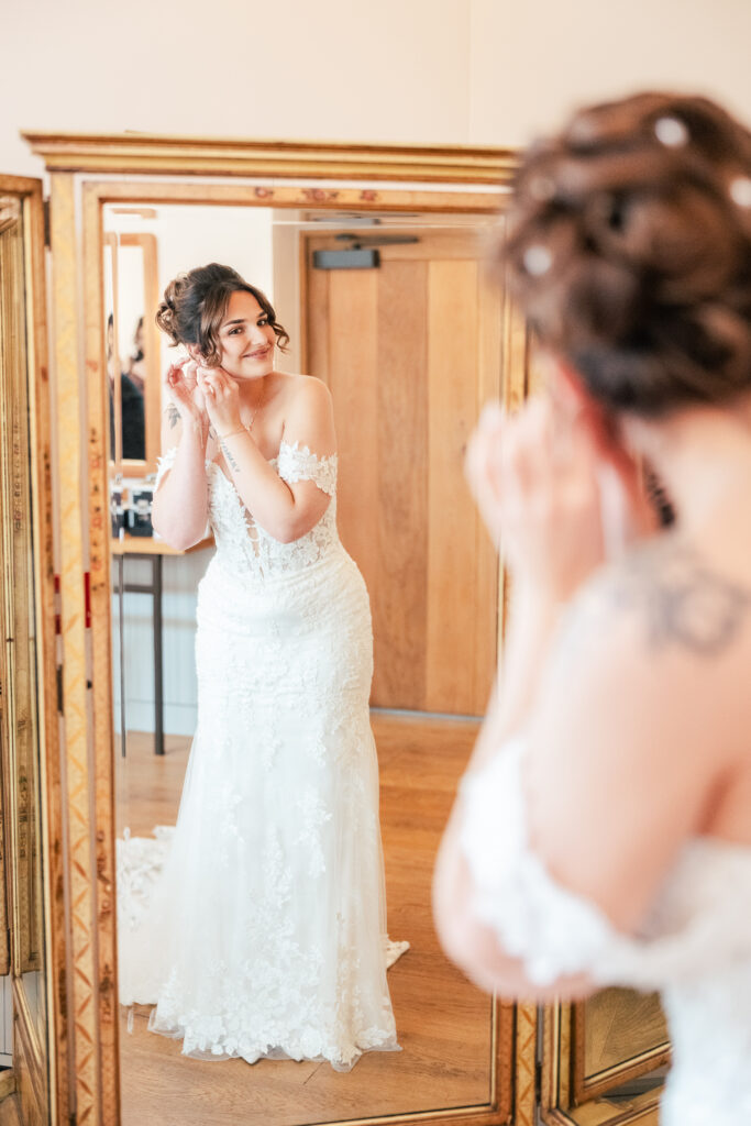 A bride in a white lace dress smiles whilst putting on earrings, standing in front of a large mirror with a wooden frame, reflecting her image in a bright room.