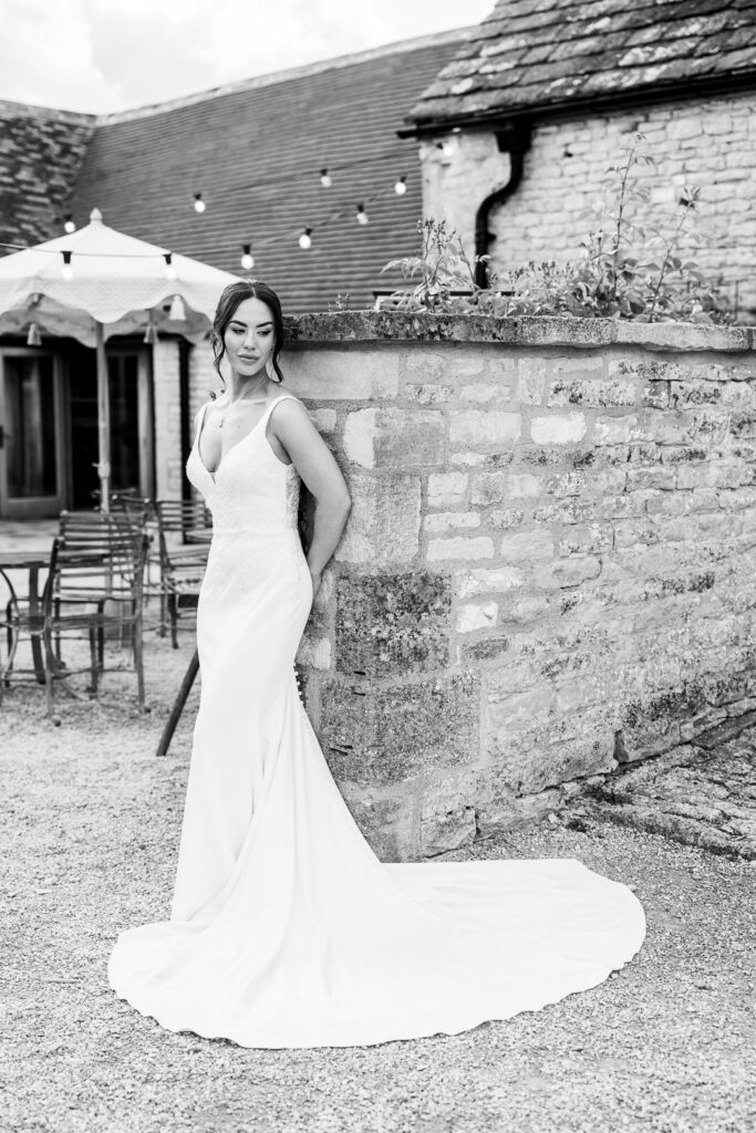 A woman in a long, elegant wedding dress stands against a stone wall outdoors, with a rustic building and empty chairs in the background. The image is in black and white.