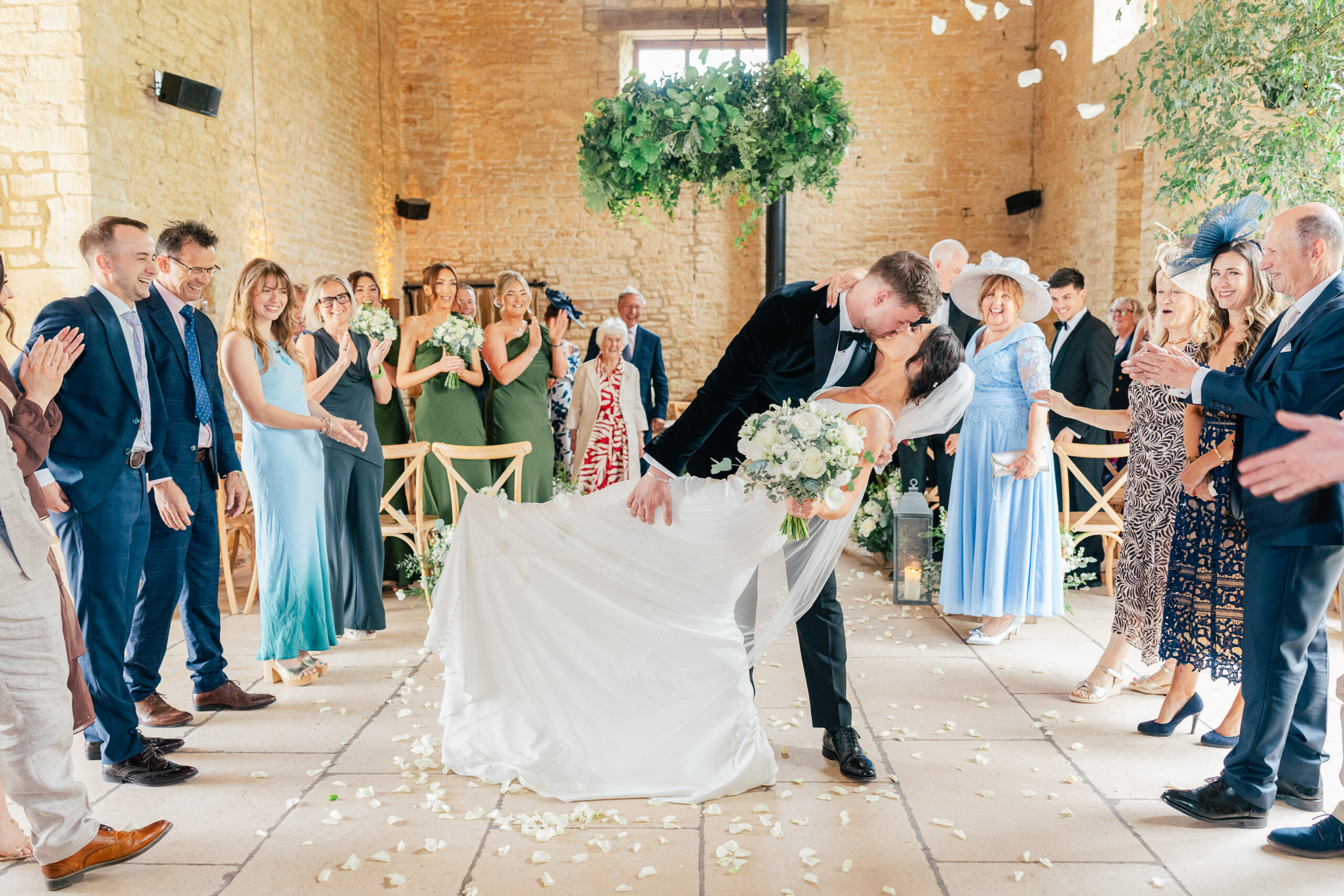 A bride and groom share a kiss as the groom dips the bride in the aisle, surrounded by cheering and smiling wedding guests in a bright, rustic venue decorated with flowers and greenery.