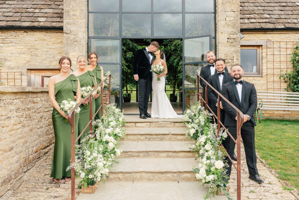 A bride and groom kiss at the top of stone steps outside a rustic venue, flanked by bridesmaids in green dresses and groomsmen in black tuxedos, with white floral arrangements lining the railings.