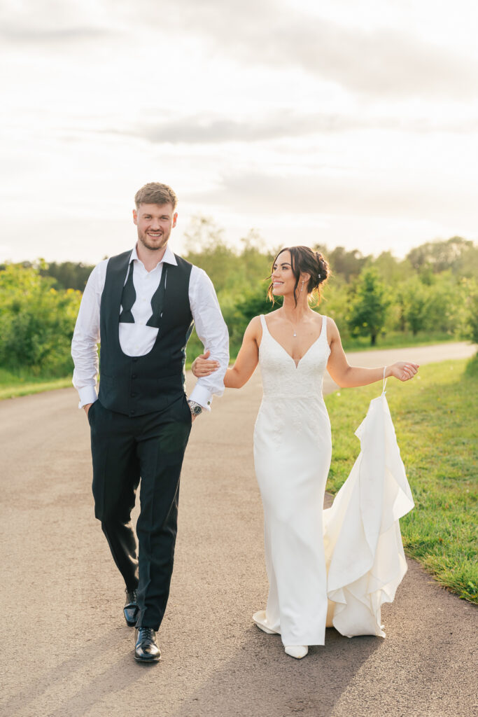 A bride in a white dress and a groom in a black suit without his jacket walk hand-in-hand on a paved path outdoors, smiling at each other, with greenery and soft sunlight in the background.
