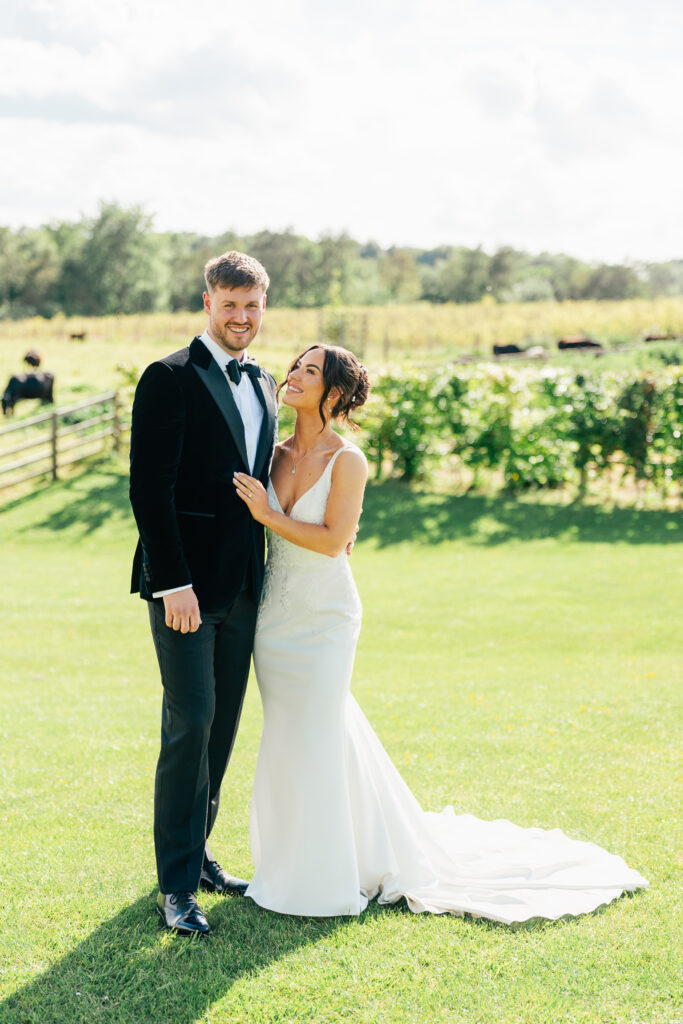 A bride in a white dress and a groom in a black tuxedo stand smiling together on a sunny lawn with green grass, trees, and a wooden fence in the background at The Old Gore in the Cotswolds.