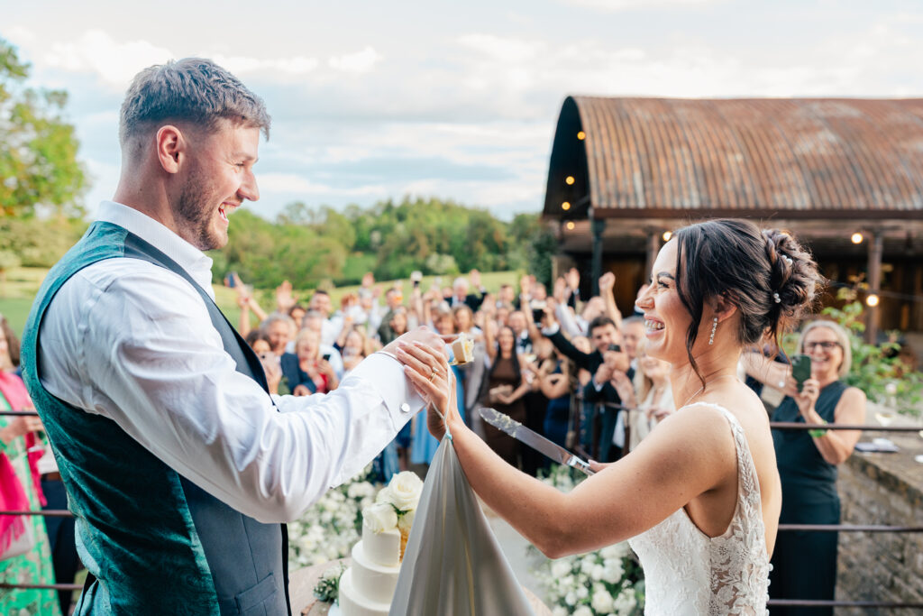 A bride and groom, smiling and holding a knife together, cut their wedding cake outdoors as a crowd of guests behind them cheer and celebrate under a partly cloudy sky near a rustic barnet the Old Ogre in the Cotswolds.