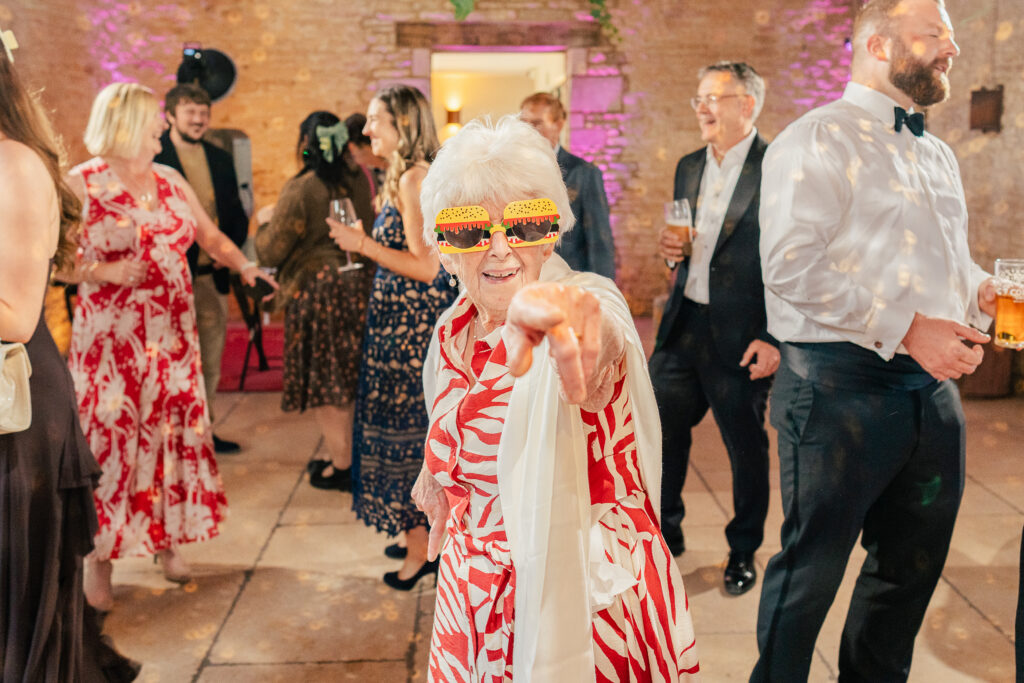 An elderly woman wearing colourful sunglasses and a red-and-white dress points at the camera, smiling, whilst people around her laugh and dance at a lively indoor party at the Old Gore in the Cotswolds.
